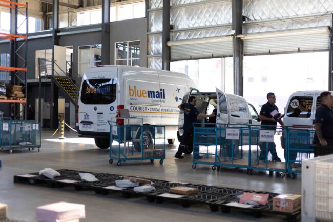 Operativa logística de Blue Mail en centro de distribución Implementación de ISO 9001 en empresas de logística: operativa del centro logístico de Blue Mail con vehículos y personal gestionando paquetería.
